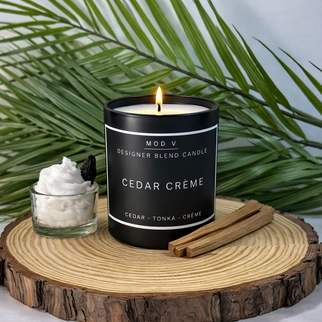 Candle labeled 'Cedar Crème' with a small bowl of cream and cinnamon sticks on a wooden surface with green leaves in the background.