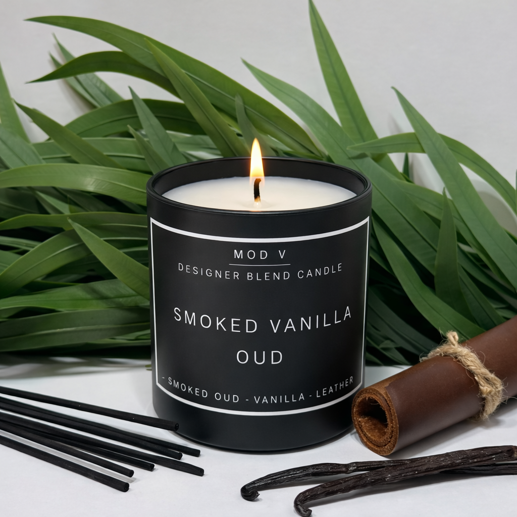 Smoked Vanilla Oud candle with packaging and vanilla beans on a white surface with green leaves in the background