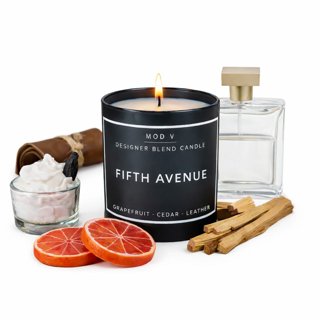 Candle labeled 'Fifth Avenue' with a lit wick, surrounded by grapefruit slices, cinnamon sticks, and a small container of white cream.