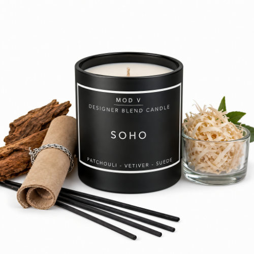 A black Mod V designer blend candle labeled 'SOHO' with patchouli, vetiver, and suede notes, styled with black straws, rolled brown paper, dried bark, and a small glass bowl of shredded natural filler on a white background.